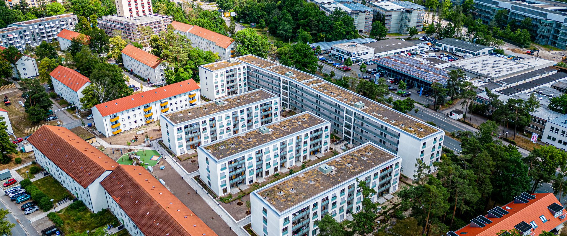  Luftaufnahme einer Wohnsiedlung mit mehreren modernen, weißen Mehrfamilienhäusern, die von älteren Gebäuden mit roten Dächern und viel Grün umgeben sind. Im Hintergrund ist ein größeres Stadtgebiet mit weiteren Gebäuden und Wäldern zu sehen.
