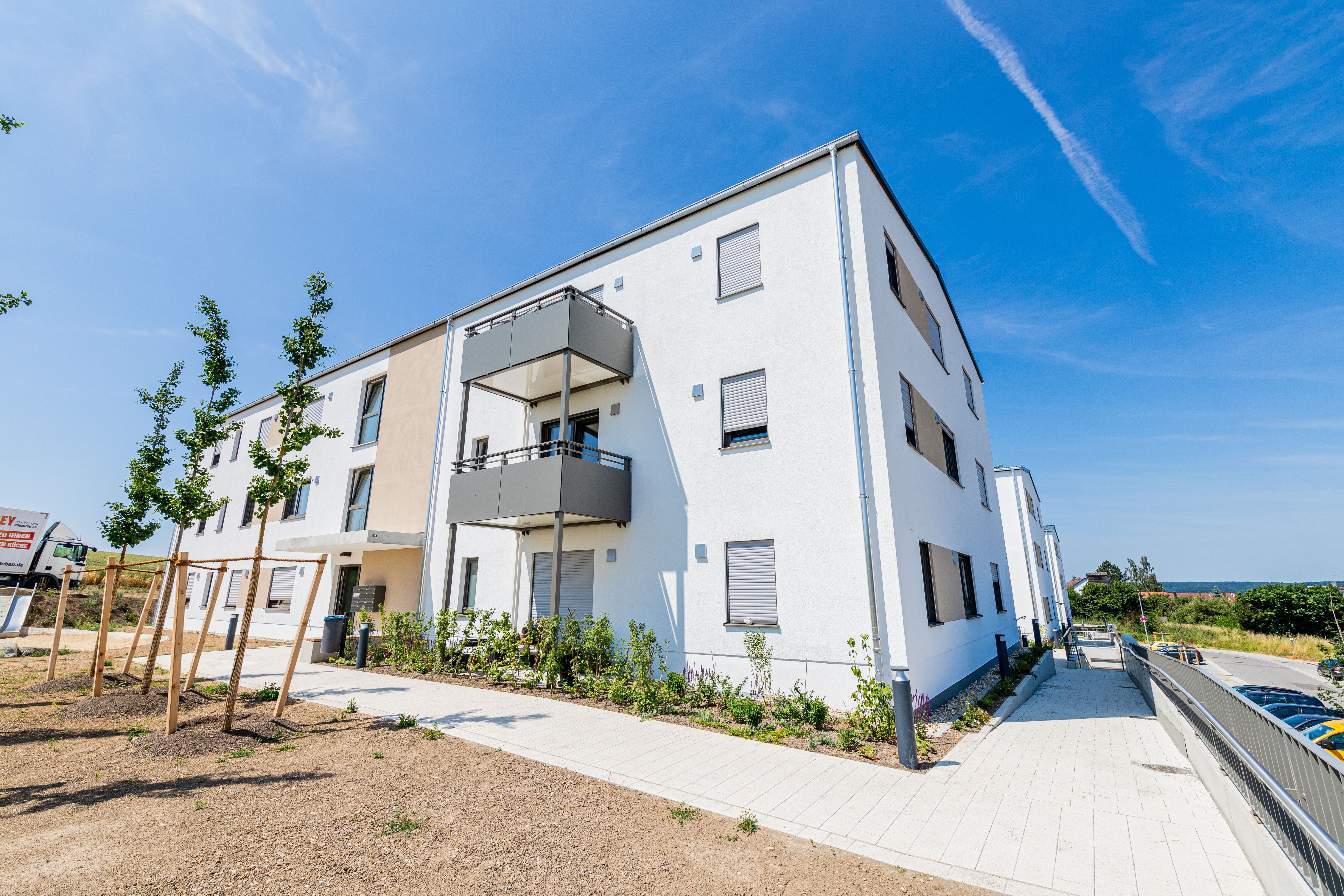 Modernes, weißes Mehrfamilienhaus mit dunklen Balkonen, davor Gehweg und junge Bäume.