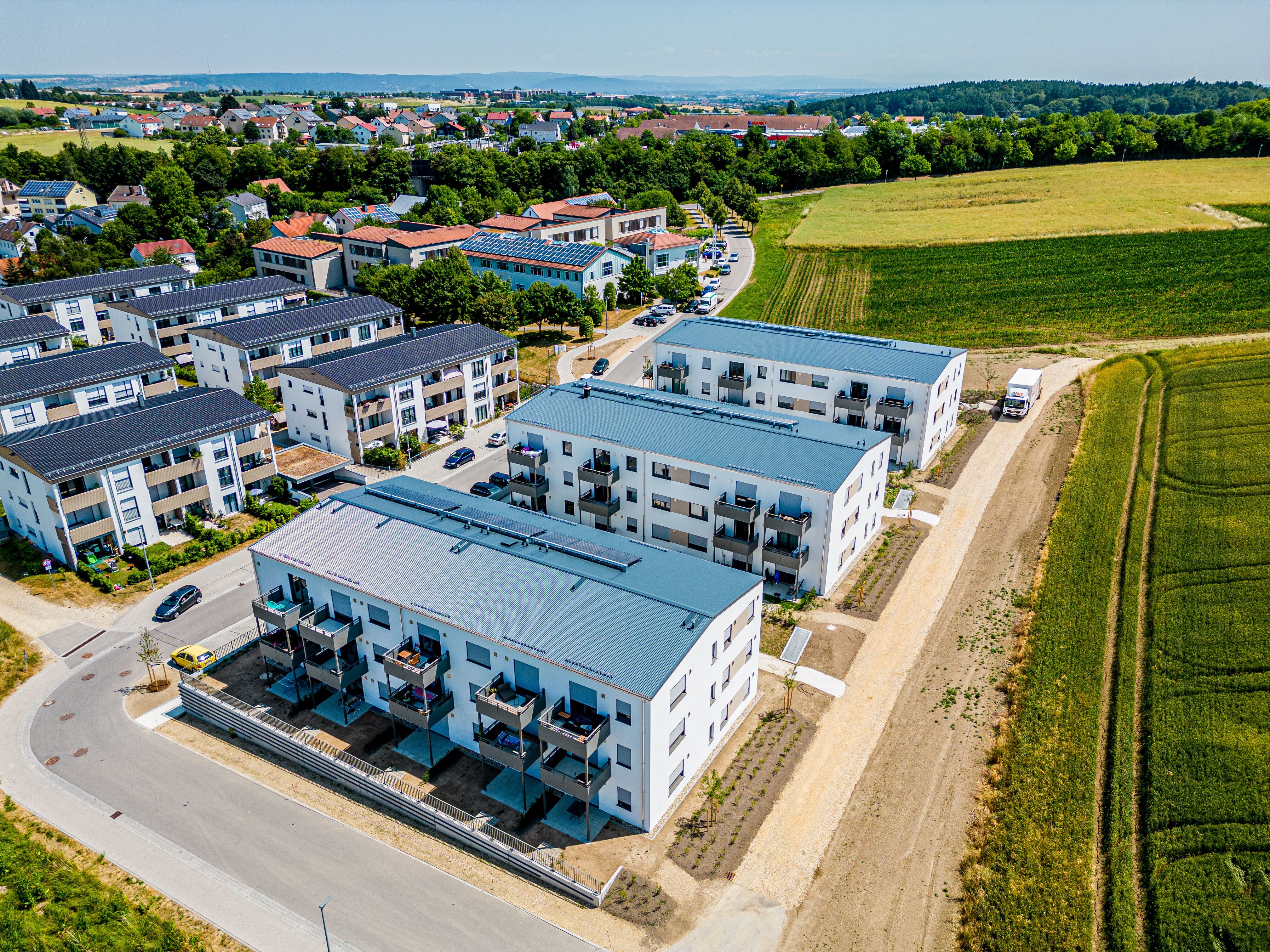  Luftaufnahme in Regensburg mit mehreren modernen, weißen Mehrfamilienhäusern mit grauen Dächern und Balkonen. Die Gebäude sind entlang einer Straße angeordnet, dahinter erstrecken sich grüne Felder und weitere Wohnhäuser in der Ferne.