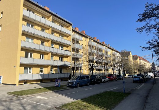 Das Quartier Rotwand in München, eine mehrstöckige, gelbe Wohnanlage mit langen Balkonreihen an einer ruhigen befahrenen Straße mit parkenden Autos unter blauem Himmel.