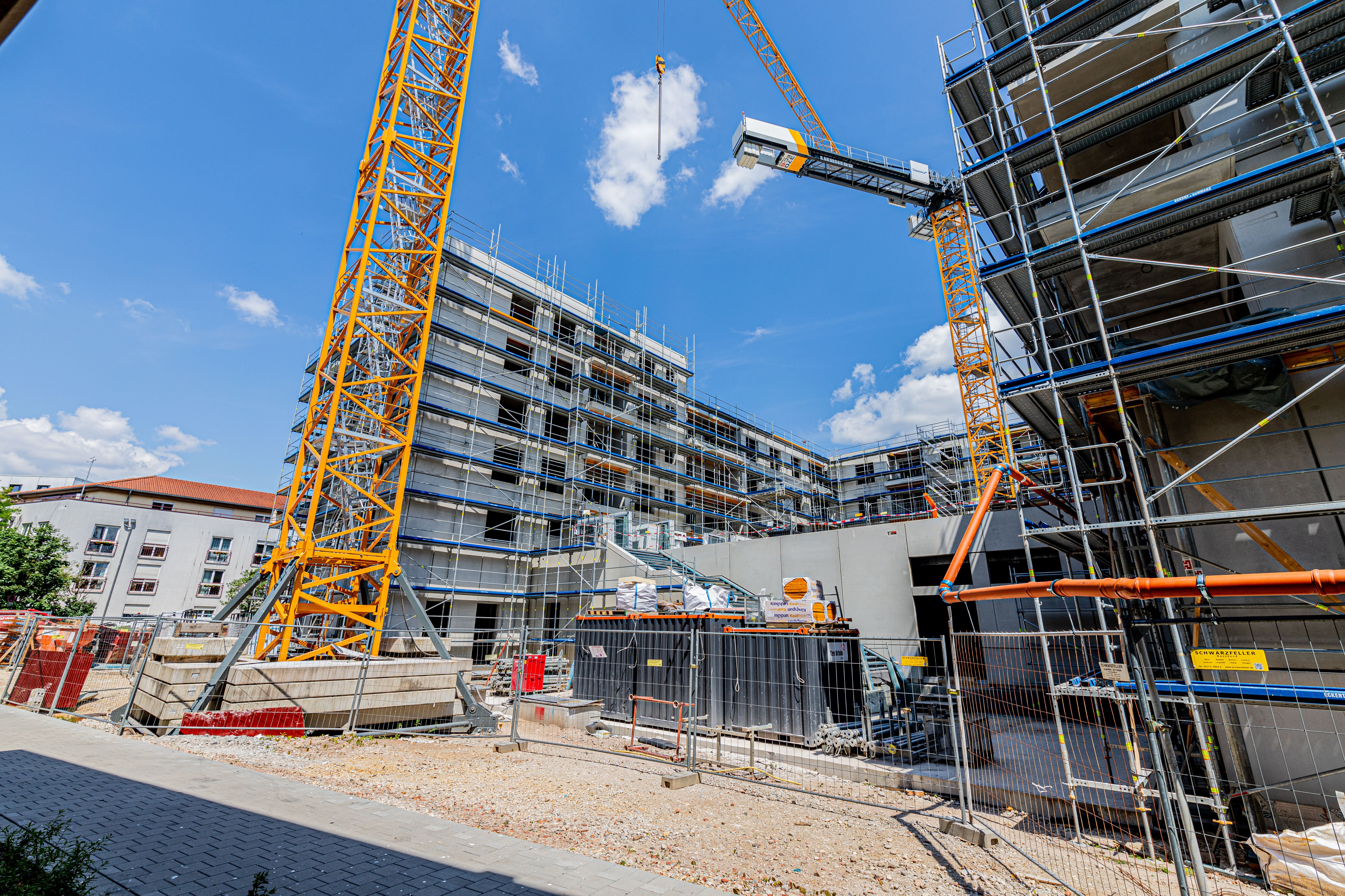 Baustelle mit mehreren eingerüsteten Gebäuden und zwei gelben Baukränen. Im Vordergrund sind Bauzäune und Materialien.