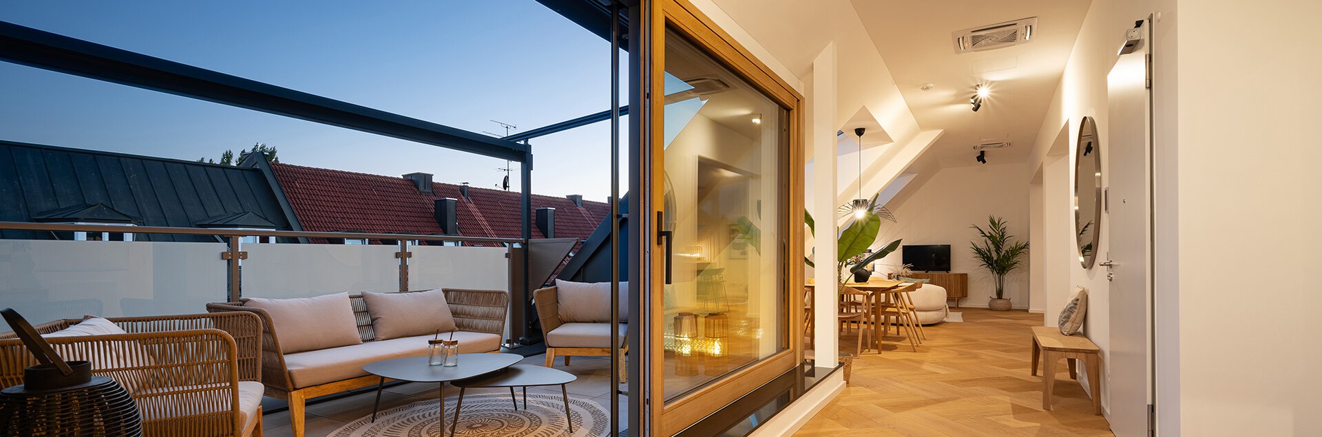 Wohnraum und Balkonanblick der Dachgeschosswohnung des Kaufobjekts Bohome. Beleuchtet und gemütlich eingerichtet mit Balkonmöbeln, Esstisch, Sofa, Fernseher und Pflanzen